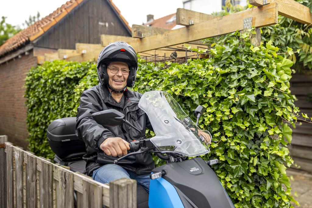Gerard zit op zijn e-scooter en is klaar voor vertrek. Hij lacht in de camera.