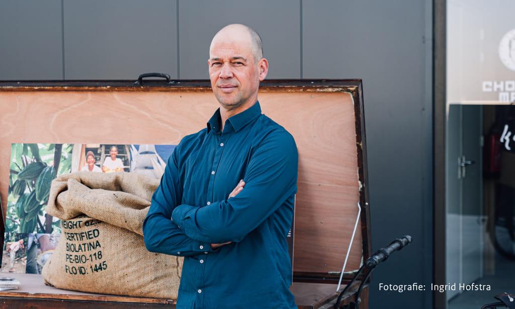 Portret van een van de chocolatemakers voor een grote, open kist waarin een zak chocoladebonen ligt.