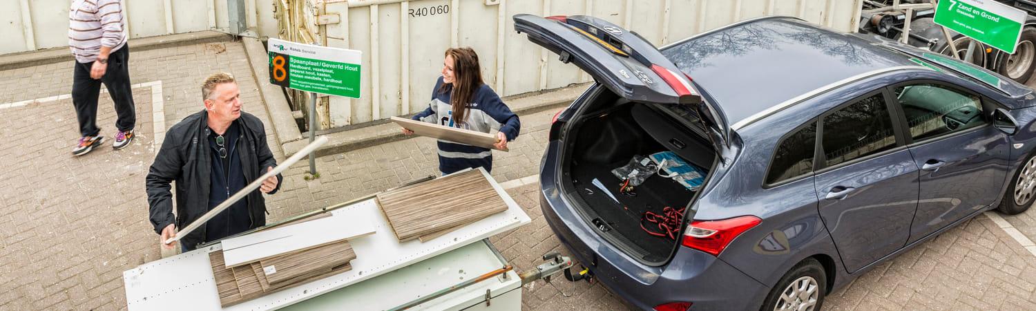 Foto van een man een vrouw die met een auto door een afvalstation rijden. Ze gooien houten planken weg die in hun aanhangwagen liggen.