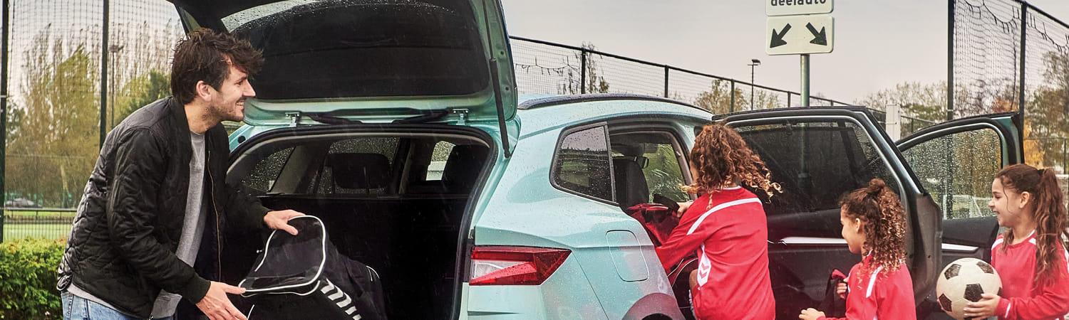 Een vader haalt zijn dochters op van voetbaltraining met een deelauto. Hij legt de sporttas in de kofferbak van de blauwe deelauto, terwijl de dochters op de achterbank gaan zitten.