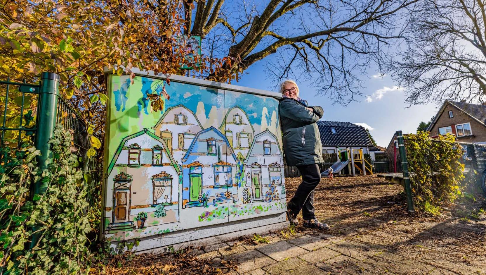 Een elektriciteitshuisje is vrolijk beschilderd met een tekening van verschillende, kleurrijke huisjes. Een vrouw poseert naar het huisje.