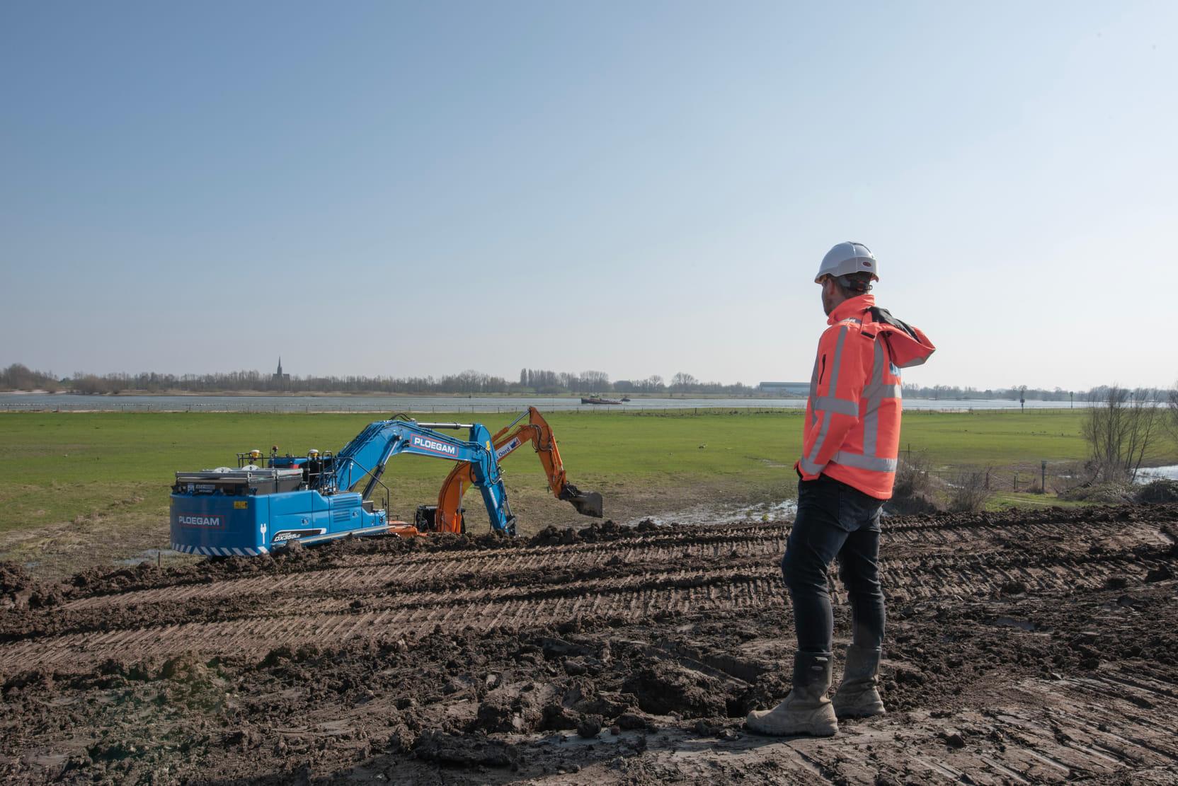 Een man in een oranje vestje en helm kijkt naar een graafmachine die aan het werk is.