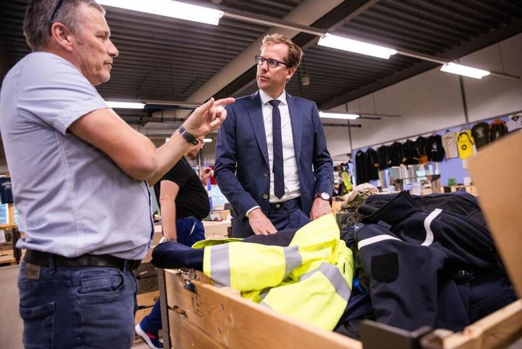De staatssecretaris krijgt uitleg van een medewerker over hoe kleding wordt hergebruikt. Ze staan bij een bak met uniforms.