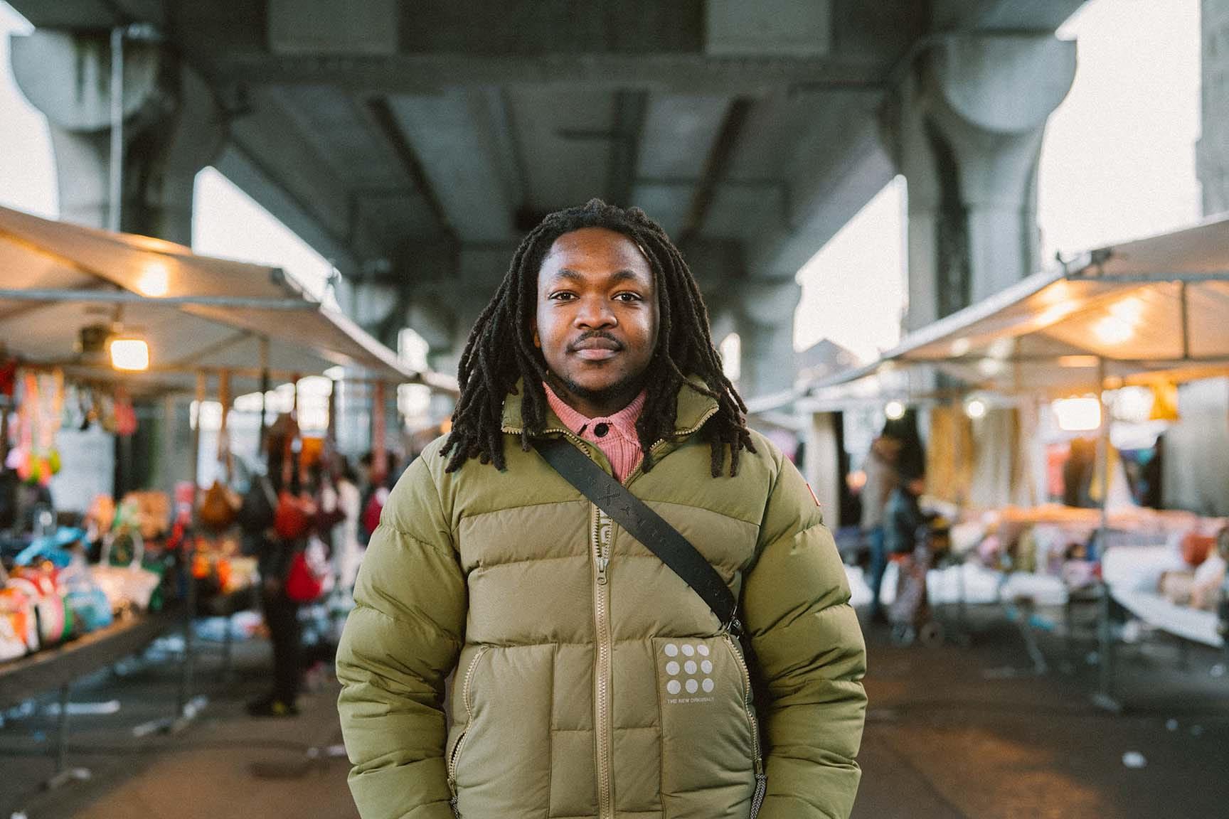 Een man in een groene jas kijkt de camera in. Hij staat voor een markt met kraampjes.