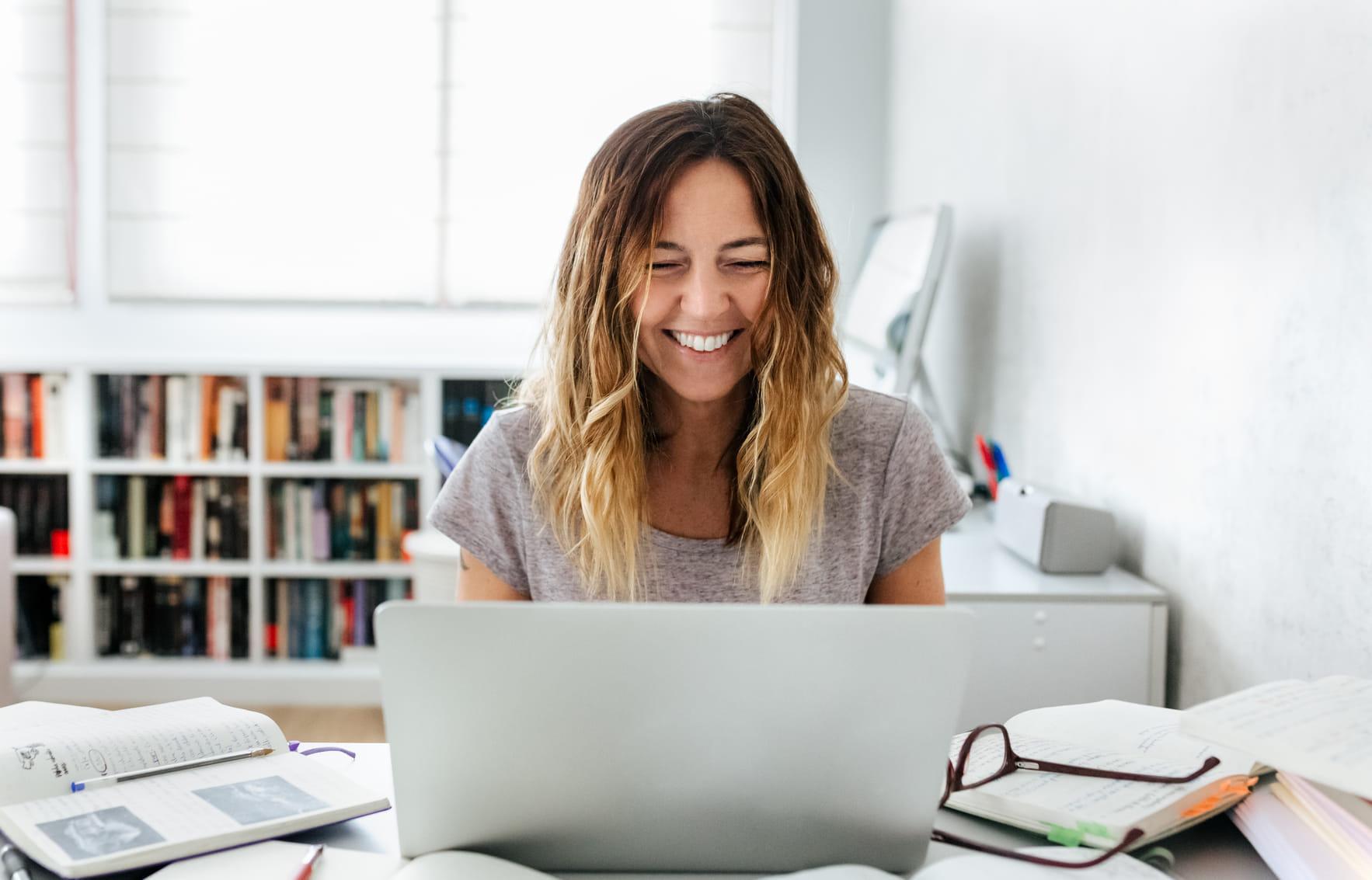 Vrouw is thuis aan het werk in een lichte ruimte. Ze kijkt lachend naar het laptopscherm.