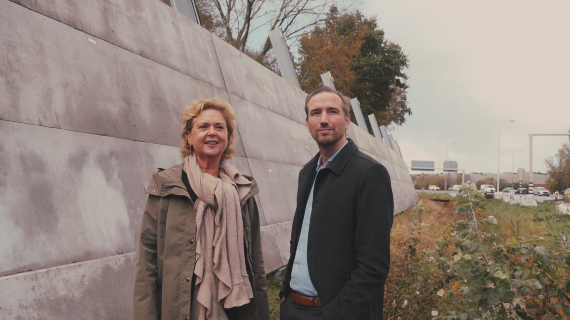 Inwoner en medewerker van de gemeente staan naast de geluidswering-schermen langs de snelweg.