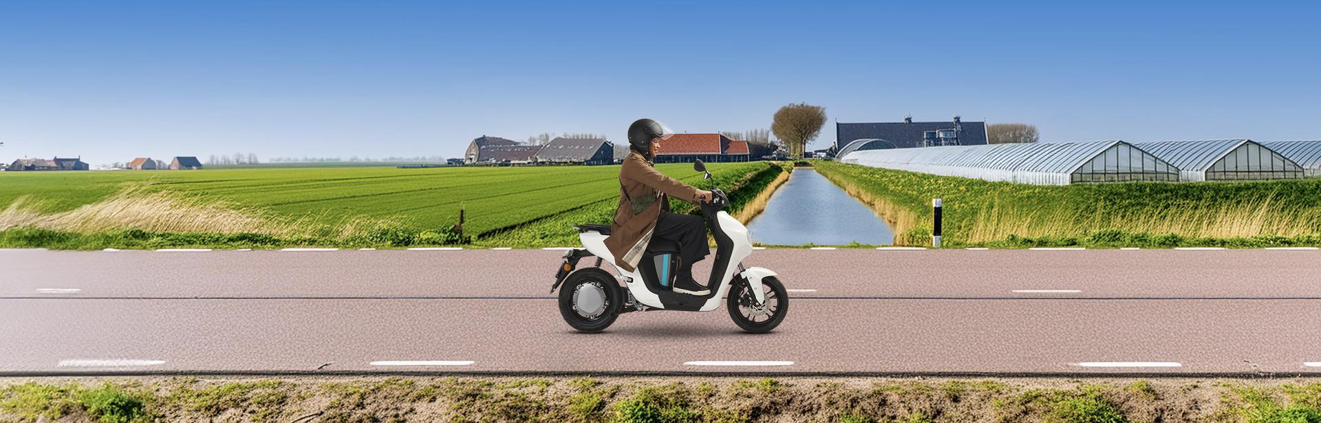 Een vrouw zit op een e-scooter en rijdt door de polder op een weg. Op de achtergrond zie je kassen, een huis en boerderij.