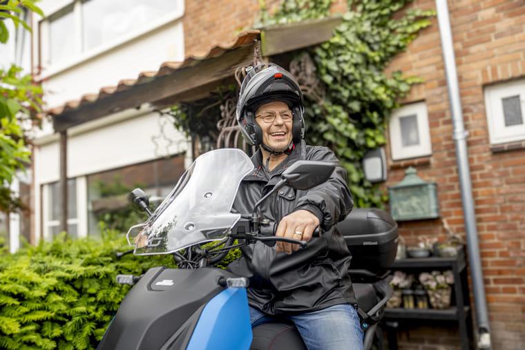 Gerard zit op zijn e-scooter en is klaar voor vertrek. Hij lacht en kijkt naar rechts.