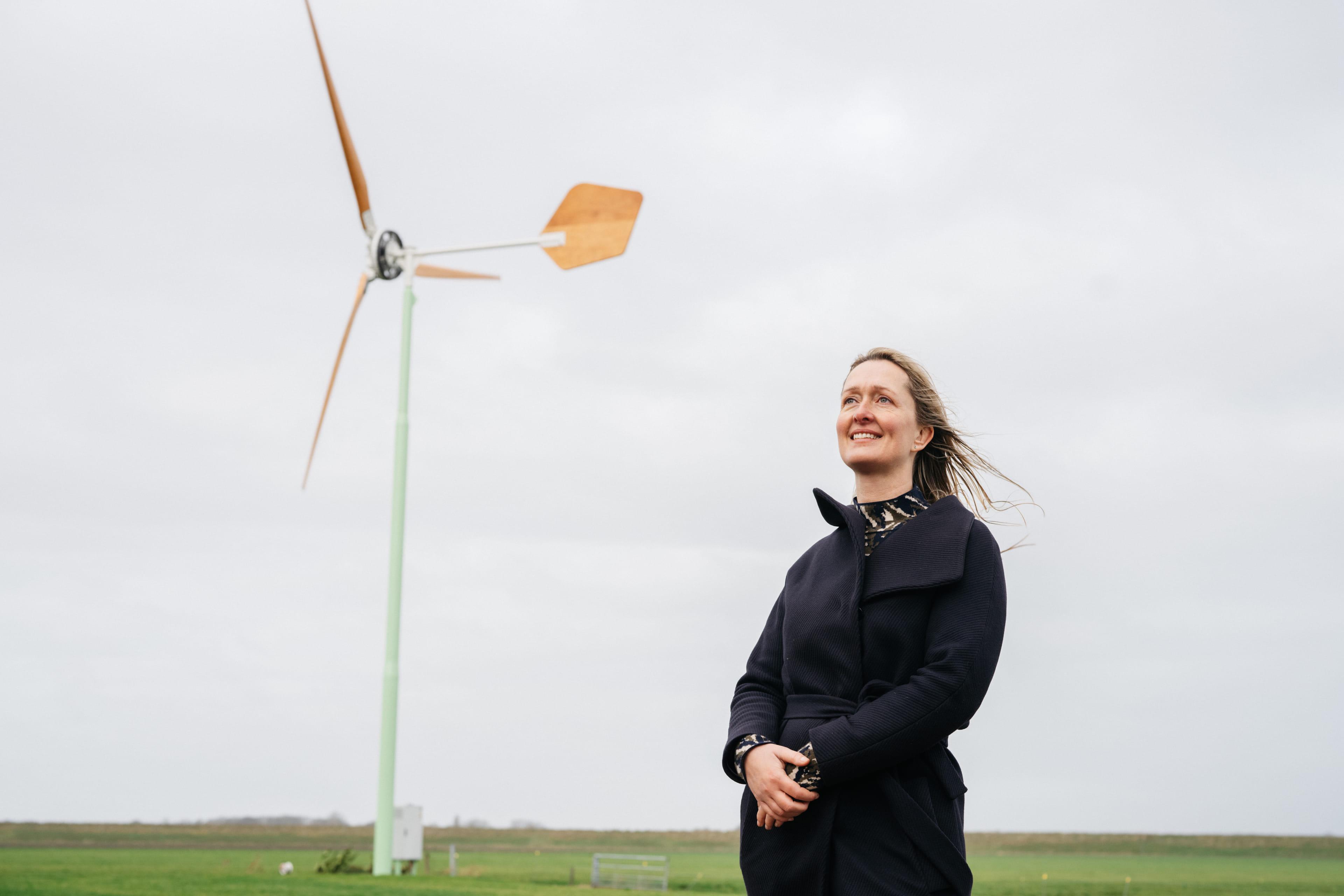 Vrouw in zwarte jas staat voor een windmolen en lacht.