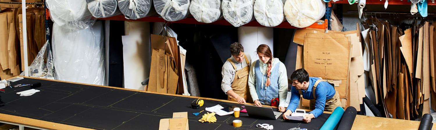 Drie mensen staan aan een ontwerptafel in een stoffenfabriek en hebben werkoverleg.