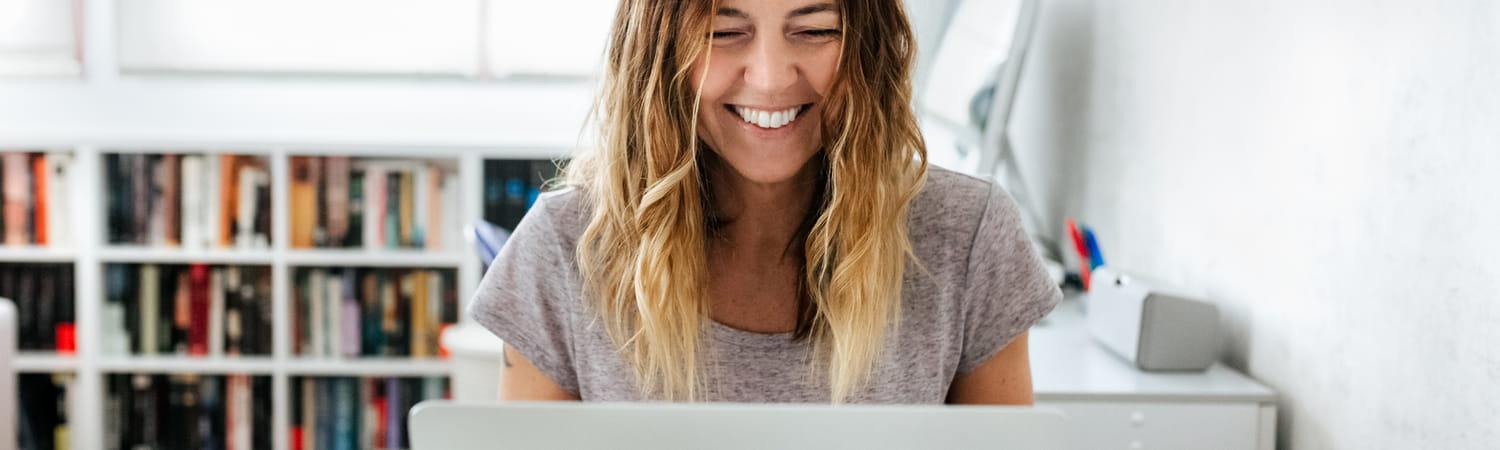 Vrouw is thuis aan het werk in een lichte ruimte. Ze kijkt lachend naar het laptopscherm.