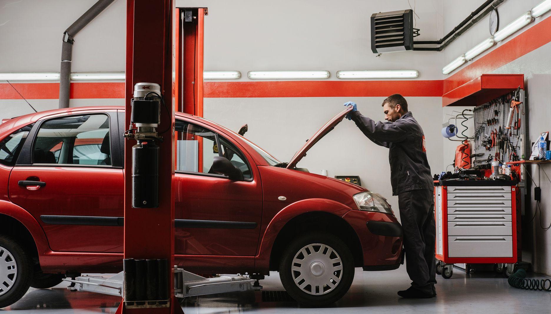 Een automonteur in een garage maakt de motorklep van een rode auto open en kijkt omlaag.