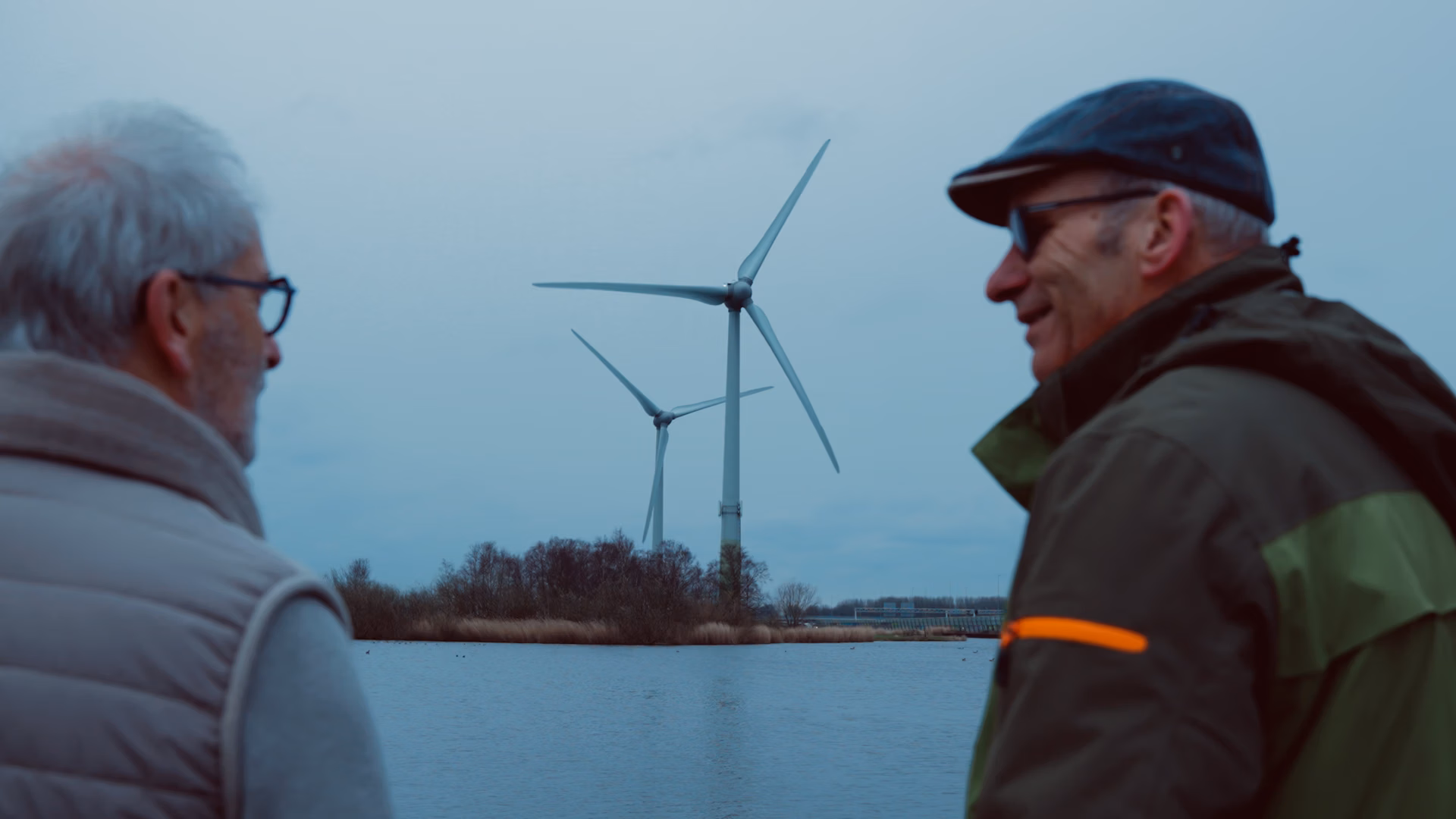 Rob Boerée en buurtbewoner kijken uit op een plas water met daarachter een stuk grond met de Watergeuzen-windmolens.